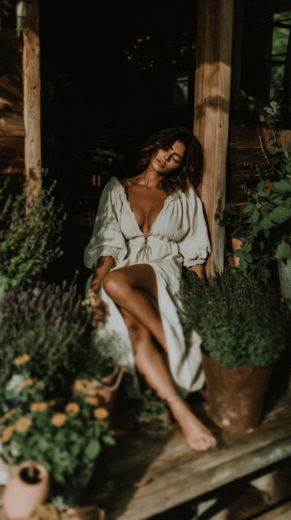 Woman in a white dress sitting on a rustic porch surrounded by potted plants in soft, natural light.