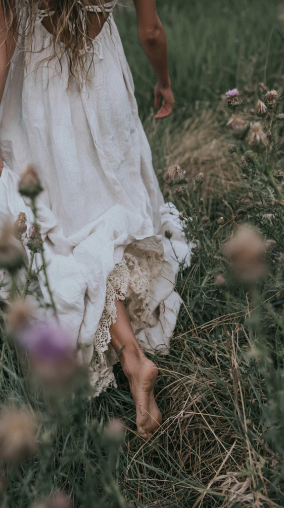 spring2 Barefoot woman in boho white linen dress walking through wildflower meadow, lace hem brushing grass